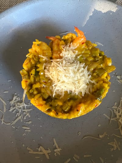 A top-down view shows a portion of saffron risotto on a gray plate. The risotto, a vibrant yellow hue, appears to contain diced green vegetables and two shrimp. It's topped with a mound of shredded parmesan cheese. Stray shreds of cheese are scattered across the plate's surface around the risotto. The plate itself has a slight sheen and hints of gray speckling.