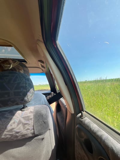 This media file captures an interior view from a vehicle, looking upwards and outwards through an open sunroof. The scene appears to be set during the daytime, under a clear blue sky, suggesting fair weather.
The primary subjects are two individuals inside the car. One person, partially visible in the foreground, has blonde hair tied back in a ponytail, suggesting a young female. Another individual, wearing a grey t-shirt, is seated to the right. The interior of the car is visible, including upholstered seats with a patterned fabric, and parts of the car's ceiling and door frame. The open sunroof reveals a landscape of a green field, possibly crops, stretching towards the horizon under the bright sky. This view provides a strong clue about the location, suggesting a rural or agricultural setting.
The image appears to be taken from within the car, possibly by one of the occupants, as they are looking out at the passing scenery. The overall mood conveyed is one of travel and observation, with a sense of openness due to the sunroof.
There is no visible text in the media file. The location context provided, "Слабодкаўскі сельскі Савет, Belarus," further solidifies the rural setting and suggests a specific region within Belarus.
