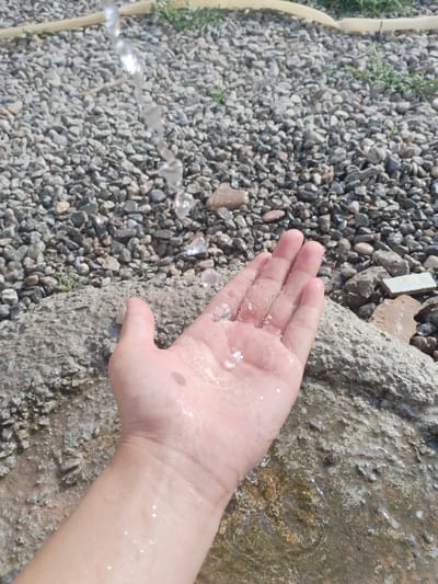 The media file shows a close-up view of a person's hand cupped to catch a stream of water. The hand is pale and appears wet, with water droplets visible on the skin and in the palm. The water is falling from above, creating a transparent stream.
The background consists of a surface covered in small, gray pebbles, with some patches of green vegetation. A large, rough, gray rock formation is also visible in the foreground, with water pooling on it. In the upper portion of the image, a yellow hose or pipe is partially visible, from which the water appears to be originating. The overall scene suggests an outdoor setting on a sunny day, likely in a natural or semi-natural environment.
The activity depicted is the act of collecting or receiving water in one's hand. The focus on the hand and the water creates a sense of simplicity and perhaps a moment of refreshment or play. There are no visible emotions conveyed due to the framing of the shot. The time of day is suggested to be daytime due to the lighting, and the weather appears to be clear. Location cues like the pebbles and rock formation are common in many outdoor areas. There is no visible text in the media file.