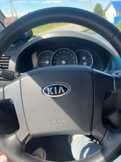 The image shows the interior of a Kia vehicle, viewed from the driver's perspective. The steering wheel, with the Kia logo prominently displayed, is in the foreground. The dashboard features a speedometer and tachometer, with a bright sunny sky visible through the windshield. Outside the car, a rural setting with houses and trees can be seen, suggesting the car is driving on a road near a residential area. The overall impression is that of a casual drive on a clear day in a suburban or rural environment.