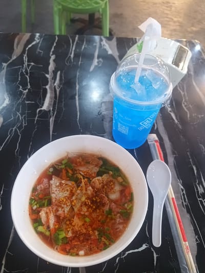 On a black marble-patterned table, likely outdoors, a bowl of Thai boat noodles sits next to a plastic cup filled with a bright blue beverage containing ice cubes. Beside the bowl are a white plastic spoon and chopsticks still in their wrapper. In the background, the blurred image of a green plastic chair is visible. Based on the "ou_ton" text on the drink cup, it is likely the photo was taken at the restaurant "Ou Ton อาหารคุณต้น" in Thap Sakae, Thailand.