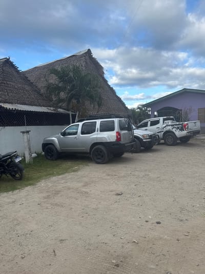 The image displays an outdoor scene in La Ceiba, Honduras, under a partly cloudy sky. The foreground consists of an unpaved dirt and gravel surface showing tire tracks.
On the far left, partially visible on a patch of green grass, is a black motorcycle with a Honduran license plate displaying "AG 0223". Adjacent to the grass, a silver Nissan Xterra SUV is parked, featuring black wheels, a black roof rack, and a black aftermarket rear bumper with a tire mount. Behind the Xterra, a white pickup truck, possibly a Toyota Tacoma, is partially visible. To its right, a white Nissan Frontier pickup truck with distinctive black and white graphical decals on its bed is parked near a building.
In the mid-ground, two structures with large, steeply pitched, natural thatched roofs are prominent. The leftmost thatched building has a white lower wall with dark lattice paneling. A palm tree stands between these two thatched structures. To the right, a light purple building with a flat, light-colored roof and a dark brown panel (likely a door or shutter) is visible. A thin, dark wire extends horizontally across the sky in the upper mid-ground. No individuals are present in the scene. The architectural style, particularly the thatched roofs, is characteristic of a coastal or tropical resort/restaurant area within La Ceiba.