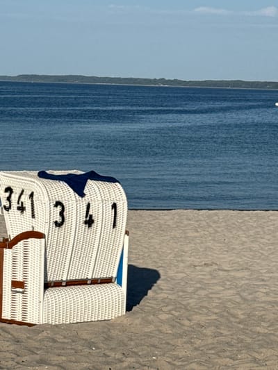 A white woven beach chair with the numbers "341 341" printed in black is positioned on a sandy beach. A dark blue fabric is draped over the top of the chair. The chair casts a shadow on the sand to its right. In the background, a calm blue ocean stretches to the horizon, where a tree-lined landmass is visible under a clear, light blue sky. The scene appears to be located on the Baltic Sea coast in Eckernförde, Germany, as indicated by the typical beach chair design and the coastal landscape.