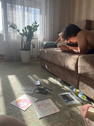 A room interior in Uryupinsk, Russia, is depicted with a person lying on a brown sofa. The person has dark, curly hair and is wearing blue shorts. On the floor, which is covered with a patterned rug, are drawing materials: markers, drawings of cats and a character with pink hair, a water bottle labeled "Active", and a case for markers. A ZZ plant in a white pot is positioned in front of a window covered with blinds. A pink slipper is partially visible on the rug. The scene is illuminated by sunlight.
