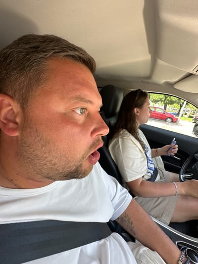 The image shows two people in the front seats of a car. The person in the foreground, a man, is looking up and to the left, with his mouth slightly open and his tongue visible. He has short, graying hair and stubble on his face. He is wearing a white t-shirt and a seatbelt.

In the background, a woman is lying back in the passenger seat, looking up and to the right. She has long, dark brown hair and is wearing a light-colored t-shirt with a striped pattern on the shoulders. She is holding a blue object in her right hand, possibly an e-cigarette. She is also wearing bracelets on her left wrist. Outside the car window, trees and a red car are visible, suggesting it is daytime and likely sunny. The interior of the car appears to be a modern vehicle, with leather seats and a steering wheel visible. The scene suggests a relaxed or possibly tired state for both individuals, perhaps during a road trip. There is no visible text in the image.