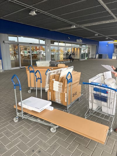 The scene is an outdoor paved area in front of an IKEA store in Sesto Fiorentino, Italy. The building facade features a prominent blue band above automatic glass doors and a dark grey corrugated ceiling. The word "Uscita" (Italian for "Exit") is visible above one set of doors.
In the foreground and midground, four trolleys are loaded with items. A long, flatbed trolley in the immediate foreground carries a large, flat, brown cardboard package and a smaller white box labeled "RINGSTA." Behind it, two similar but shorter flatbed trolleys are stacked with numerous brown cardboard boxes, many bearing the IKEA logo, indicative of flat-pack furniture. One of these trolleys also holds a white plastic basket. To the right, a standard metal shopping cart with blue handles is filled with various white and metallic household items, including a waste bin and possibly a lighting fixture.
On the far right, partially visible, are the lower arms and hands of two individuals; one holds a clipboard with papers. In the background, two individuals are seated on a wooden bench located in front of the store's glass entrance/exit. Reflections in the glass doors show a white Jeep and other indistinct figures in a parking area beyond. The paving is composed of grey bricks arranged in a herringbone pattern.