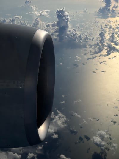 The image shows the engine of an aircraft in flight. The view is from inside the aircraft, looking out at the engine nacelle, with a backdrop of clouds and possibly the ocean reflecting sunlight. There is no indication that the image was taken in Taoyuan City, Taiwan.