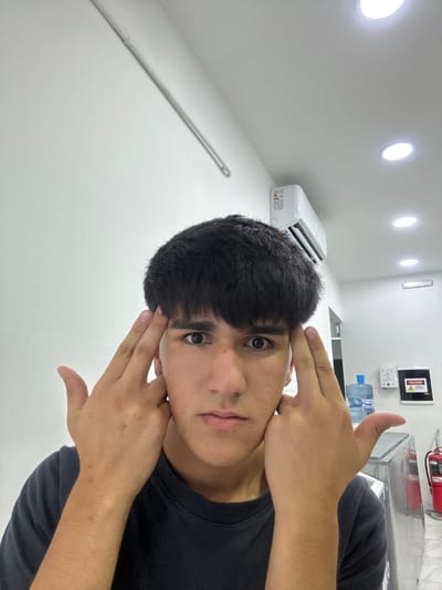 A person with short, dark hair and light skin makes a hand gesture near their head. The person wears a dark shirt and faces the camera at an angle. The background is a white wall with multiple ceiling lights, an air conditioning unit, and a water bottle on a counter. A fire extinguisher is also visible on the upper-right. The image is slightly rotated.