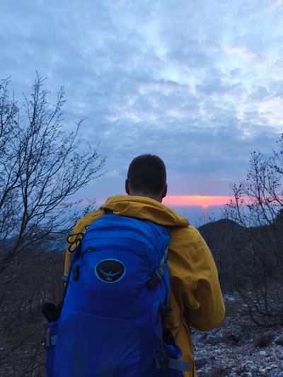 A single individual is observed from behind, facing an orange-red sunset over a distant horizon. The scene is located in Mali Mikulići, Montenegro. The person wears a yellow hooded jacket and a royal blue Osprey hiking backpack with a visible avian logo. The sky above is largely covered with broken, light blue and grey clouds. Sparse, bare-branched trees and shrubs frame the scene on either side, with the ground beneath appearing rocky and sparsely vegetated. The person is stationary, observing the sunset.