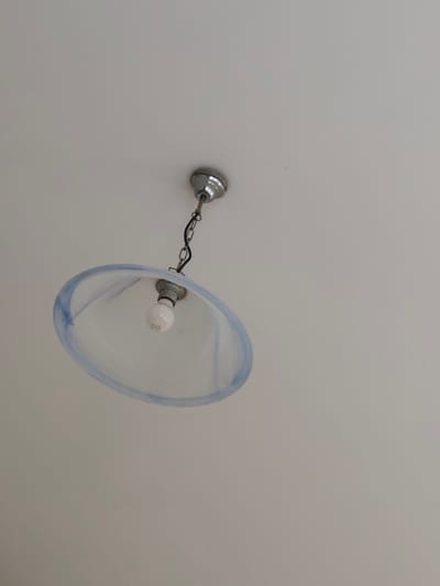 This media file shows a close-up, low-angle shot of a hanging light fixture against a plain, light-colored ceiling. The fixture consists of a chrome-colored ceiling mount, from which a black chain suspends a light blue and white glass lampshade. Inside the lampshade, a single, white, light bulb is visible. The lampshade has a somewhat oval or bowl shape, with a translucent quality and subtle swirling patterns of blue marbling. The overall scene is simple and domestic, with no people or significant activity taking place. The lighting suggests it could be daytime, as the ceiling is brightly illuminated, but the artificial light from the bulb is also present. There are no visible text elements in the image. The setting appears to be an indoor environment, possibly a home or a simple commercial space.