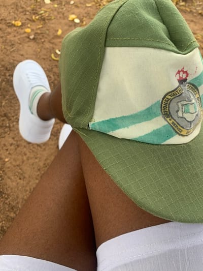 The image captures a close-up, downward-angled view of an individual's lower body, situated outdoors in Kubi A, Nigeria. A dark olive green and off-white cap, featuring the emblem of the Nigerian National Youth Service Corps (NYSC) with a crown and a map of Nigeria, rests on the person's lap. The cap's design includes two horizontal teal stripes on its off-white central band. The individual wears white shorts or trousers, white athletic socks with light green stripes, and white sneakers on the left foot. The visible skin on the legs is dark brown. The background shows reddish-brown earth scattered with dry, yellowish-brown leaves.