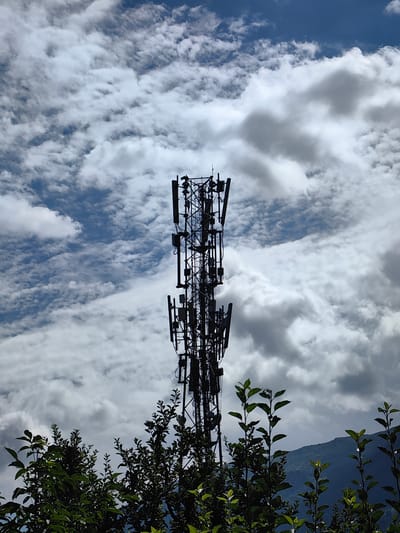 A silhouetted telecommunications tower stands tall against a dramatic sky filled with white and gray clouds. The tower is a lattice structure with numerous antennas and equipment attached to it. In the foreground, dark green foliage of trees and bushes partially obscures the base of the tower and a distant blue mountain. The image appears to be taken from a low angle, looking upwards at the tower. The location is identified as Chhyal, India. There are no people visible in the image.