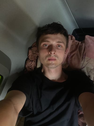 A man is lying down, facing the camera. He is wearing a black t-shirt. The background appears to be the interior of a vehicle, possibly a truck cab, with fabric upholstery visible. The man's face is in focus, and he has light-colored eyes and a fair complexion with freckles. The image is lit from above, casting shadows on his face. The overall scene suggests a candid, personal moment. There is no visible indication of the specific location within Brachterbeek, Netherlands.