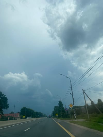 This is a photograph taken from a moving vehicle on a highway, likely approaching the city of Mironezh'e, Russia, as indicated by the presence of road signs and architecture consistent with the region. The sky is overcast with dramatic, dark storm clouds, suggesting an impending storm. The road itself is a multi-lane highway with clear lane markings, including a yellow line separating oncoming traffic and white lines delineating lanes. On the left side of the road, there is a concrete barrier and a glimpse of residential buildings with red roofs. Further along the road, several streetlights and utility poles with electrical wires are visible. Traffic signs, including a yield sign and a warning sign with an exclamation mark, are present. The right side of the road is bordered by a grassy verge and trees, with some rural structures peeking through the foliage. There are other vehicles on the highway in the distance. The overall scene depicts a transition into a potentially stormy weather environment on a major road.