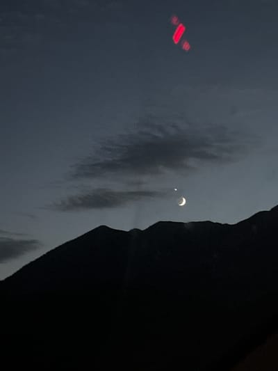 An image shows a nighttime sky over the dark silhouette of mountains. A sliver of a crescent moon is visible, accompanied by a bright point of light, possibly a planet or star. In the upper right portion of the sky, there are several blurred red lights, suggesting a lens flare or distant artificial lights. The image was taken in Farchant, Germany.