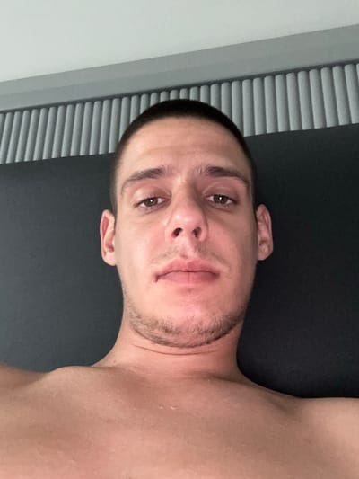 A young man is depicted in a close-up, upward-angled shot, appearing to be lying down. His head and upper torso fill the frame. He has short, dark hair and visible stubble on his chin and upper lip. His eyes, dark brown, are open and directed towards the camera with a neutral expression. His skin appears slightly flushed, possibly from heat or exertion, and there are some subtle glistening marks on his shoulders and chest, perhaps indicating sweat.

To the left side of the image, running vertically, are light-colored horizontal blinds, suggesting an indoor setting, possibly near a window. To the right and behind the man's head and body is a dark, uniform surface, which could be a bed, mat, or wall. The image has a slightly dim but even lighting. There is no visible text. The overall scene conveys a candid or casual moment indoors.