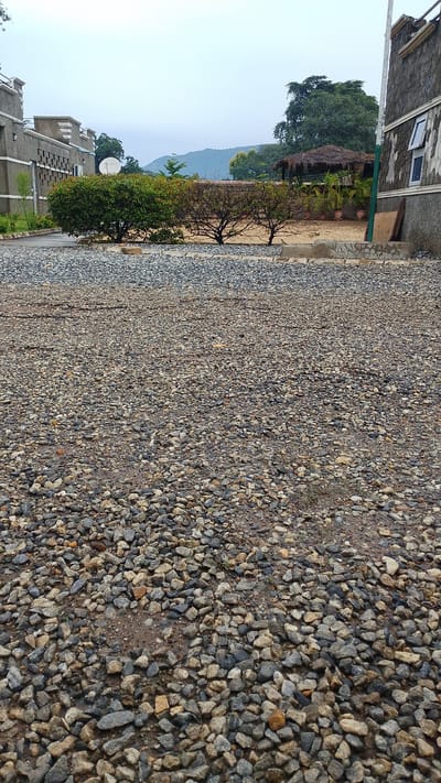 This media file depicts an outdoor scene, likely a courtyard or driveway, covered in gravel. In the background, there is a building with a grey exterior and decorative elements. Adjacent to it, a lush green bush and some trees with bare branches are visible. Further in the distance, a thatched-roof structure and more trees can be seen, with a hazy mountain range forming the backdrop. The sky is overcast, suggesting a cloudy or potentially rainy day. The gravel in the foreground appears wet in places, indicating recent precipitation. There is no visible text in the image. The scene evokes a sense of a rural or semi-rural setting in Balewa, Nigeria, with an emphasis on the natural landscape and simple architecture. No people or specific activities are depicted.
