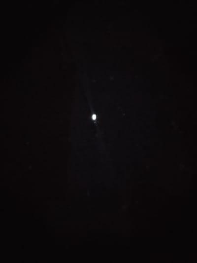 The image is a black and white photograph of a dark night sky with a single, bright celestial object, likely the moon, at the center. The object is illuminated, and a faint beam of light emanates from it, extending diagonally upwards and to the left. The rest of the image is predominantly black, suggesting the absence of significant ambient light. There are no discernible features of Rumuokwuta, Nigeria, visible in this photograph. The scene is static and devoid of any human activity or identifiable terrestrial elements.