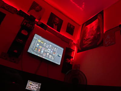 A small, red-lit room features an abstract painting, a fan, and a flat-screen TV displaying a screen of thumbnail images. A smaller screen is mounted above. Shelves are affixed to the walls. Various framed pictures adorn the walls, illuminated by a red LED strip. The overall aesthetic creates a dark, stylized, and tech-integrated space.