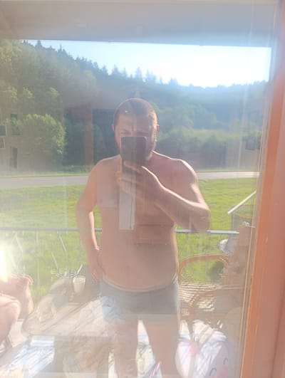 A man is taking a mirror selfie on a balcony. He is shirtless and wearing grey boxers. He is holding a smartphone up to take the picture. The balcony has a railing with a diamond pattern. A table with drinks and other objects sits on the balcony. A wicker chair is also visible. The background shows a wooded hillside and a clear blue sky. The image is taken in Berching, Germany.