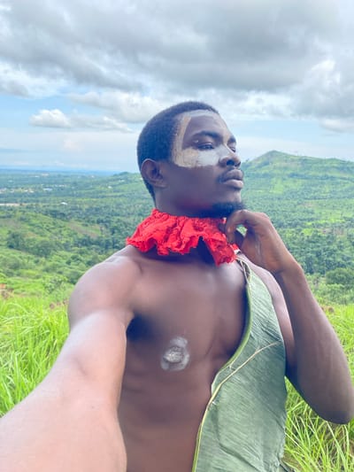 This media file is a portrait of a Black man with a red cloth around his neck and a green leaf draped over his chest. He has white markings on his face and chest. The man is looking upwards with a pensive expression. He is positioned on a grassy hillside with a lush green valley and a village in the background. The sky is cloudy. The scene appears to be set in Ufu Esioni, Nigeria. The overall mood is serene and contemplative, perhaps suggesting a connection to nature or cultural traditions.