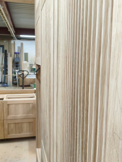 The image shows a close-up of a fluted wooden cabinet door in a workshop. The wood has a light, natural finish and a distinct grain pattern. In the background, out of focus, is a workshop environment with various tools and equipment, including a pallet jack and shelving units. The overall impression is that of a woodworking or furniture-making setting. The image does not contain any information to suggest the location is in Sofyino, Russia.