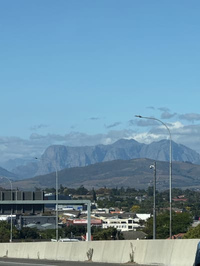 The image captures a view in Bellville, South Africa. In the background are layered mountains under a blue, slightly cloudy sky. A cityscape is situated along the base of the mountains. A grey concrete structure, possibly part of a road or bridge, is visible on the right side of the image. There are streetlights lining the road. Buildings, including commercial properties with signage like "HANDELSHUIS Auctioneers" are visible, interspersed with trees.