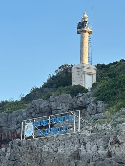 A stone lighthouse stands tall on a rocky, tree-covered hill overlooking the sea. In the foreground, a sign reads "PARK PRIRODE PLATAMUNI" with a circular logo containing a stylized depiction of a lighthouse and water. The sky is clear and blue. The scene appears to be located in Kubasi, Montenegro.