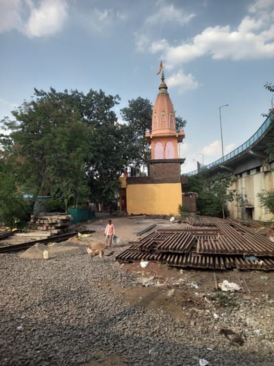 The image depicts a Hindu temple in Bhopal, India, situated near construction activities. The temple has a distinctive dome and is painted in shades of orange and yellow. It stands next to a brick building with a yellow wall. In the foreground, there's a pile of grey gravel and discarded materials, suggesting construction or renovation work. Metal beams are stacked nearby. A man in a red and white shirt is walking in the scene, carrying a construction tool. A light brown dog is standing next to him. The backdrop includes a bridge and lush green trees, with a partially cloudy sky.