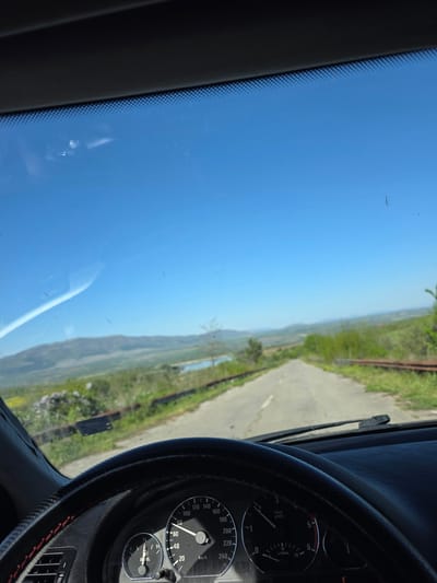 The image captures a first-person perspective from inside a car, looking out through the windshield onto a scenic rural landscape in Lelintsi, Bulgaria. The vibrant blue sky, mostly clear with a few dust specks on the glass, dominates the upper part of the frame, indicating a bright, sunny daytime. A paved road stretches into the distance, appearing to wind through rolling green hills and valleys. Lush green vegetation, including what might be trees and grass, lines the roadside and covers the distant terrain. A body of water, possibly a river or lake, is visible in the middle distance.
Inside the car, the dashboard and part of the steering wheel are visible in the foreground, primarily on the right side of the image. The steering wheel is black, textured, and features red stitching. The instrument cluster on the dashboard includes a speedometer displaying a reading of approximately 60 km/h, a tachometer showing 0 x1000 1/min (0 rpm), and a fuel gauge indicating a fuel level slightly below half, with the word "DIESEL" clearly visible. An orange warning light is illuminated on the dashboard. The windshield shows reflections and some minor dust or streaks, along with the black frit band on its left edge. The scene suggests a journey in progress or a stop along a picturesque route.