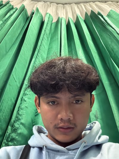 A young man with dark, curly hair and a light blue hooded sweatshirt poses for a selfie. He has a thin mustache and is looking directly at the camera. Behind him, a green curtain is draped, creating a backdrop of vertical folds. The image appears to be taken indoors, possibly in a room with decorative green fabric. There is no visual information to suggest the location is in Pekalongan, Indonesia.