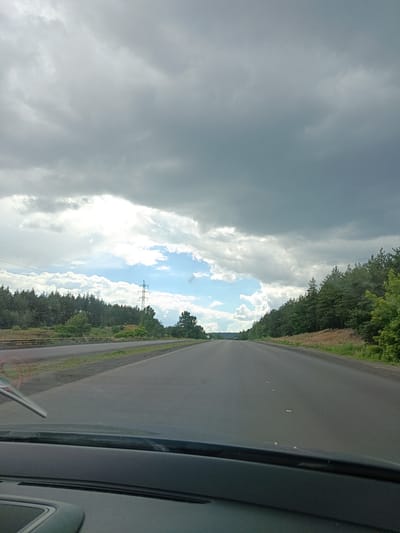 This is a forward-facing, eye-level, outdoor, full-shot photograph taken from inside a vehicle, looking out through the windshield. The scene depicts a two-lane asphalt highway in the direction of travel, with a narrow shoulder and a grassy verge on the right. A dense forest of pine trees lines the right side of the road, extending into the distance. On the left side, a similar forest is visible, separated by a row of power lines and a transmission tower. The sky is heavily overcast with dark, ominous storm clouds, but a patch of bright blue sky with scattered white clouds is visible towards the center-left. The road is empty except for a single car in the far distance. The foreground shows the dashboard of the vehicle, including a windshield wiper. The location is identified as Biloziria, Ukraine, on a highway. The time of day is likely afternoon, given the light conditions. The overall atmosphere is one of impending rain.