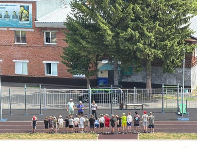 In Tashtagol, Russia, a group of children stand in a line on an outdoor athletic track, facing away from the camera. They appear to be participating in a sports activity, possibly supervised by the three men standing behind them. The men are positioned near a fence and a black car. The background features a brick building with multiple windows, large evergreen trees, and a metal roof. The overall setting suggests a school or community sports facility.
