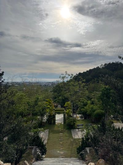 The image depicts a landscape in Liên Nghĩa, Vietnam. A pathway leads through a green, grassy area flanked by trees and shrubs. The path has stone or concrete sections. There are benches along the path. In the distance, a cityscape is visible beneath a cloudy sky. The sun is seen in the upper left corner of the image.