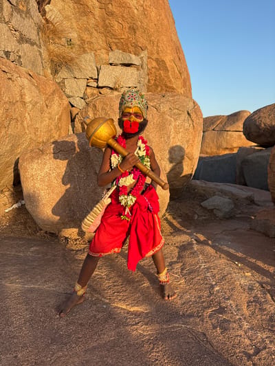 A color photograph depicts a person dressed as the Hindu deity Hanuman, situated amongst large rock formations, likely in Kamalapura, India. The individual is adorned with a headdress, facial paint including a red mouth, necklaces of flowers, and bracelets. They are clad in red clothing and holding a golden mace. The setting appears to be a natural landscape with smooth, weathered rocks and a clear sky in the background.