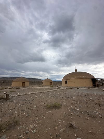 The image depicts three dome-shaped structures situated in a dry, rocky, and sparsely vegetated environment. The structures are of varying sizes, with the largest and closest one on the right featuring an extended entranceway with wooden supports. The middle and farthest structures are smaller and appear to have simple wooden doors. All structures are a uniform light brown or tan color. A wooden signpost is visible in the lower left corner. In the background, a weathered wooden fence runs horizontally, with a mountain range in the far distance under a heavily overcast sky filled with dark, dramatic clouds. The ground is covered in gravel, small stones, and patches of dry grass. This scene is located at the Dunhuang-Jiayuguan Silk Road Scenic Spot in Jiayuguan, China, representing historical dwellings or fortifications. There are no people or animals visible in the image.
