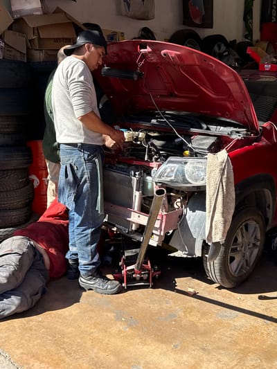 The image depicts two individuals in a garage setting, presumably in the city of Salta, Argentina. One person is lying on the ground with their legs under a red car that has its hood open. The car appears to be undergoing repair. A second person, wearing a black cap, a light grey shirt, and blue jeans, is leaning over the engine compartment, likely working on the car's engine. The surrounding environment includes a stack of tires, boxes, and tools, typical of a garage space. The lighting suggests the photo was taken during daylight hours.