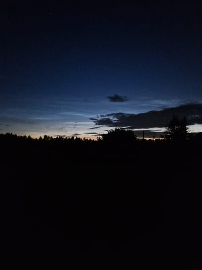A low-angle, vertical, outdoor, medium shot captures a dark, silhouetted landscape against a twilight sky. The foreground is entirely black, obscuring any details on the ground. A dense line of silhouetted trees runs across the middle ground, forming a dark barrier against the horizon. Above the trees, the sky transitions from a deep, dark blue at the top to lighter shades of blue and gray near the horizon, suggesting the last vestiges of daylight. Wispy clouds are scattered across the upper sky, and a single, faint star is visible in the top left corner. The overall impression is one of quiet dusk in a rural or semi-rural setting, possibly at the edge of Sorochkino, Russia, given the time of day and the natural elements. No people, animals, or specific man-made structures are clearly discernible due to the low light conditions and the silhouetted nature of the scene.