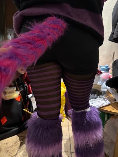 An indoor, eye-level shot captures a person from the waist down, likely showcasing a costume. The subject wears black pants or leggings, covered from the knees downwards by striking purple and black striped tights or stockings. Over these, fluffy purple leg warmers or boot covers are worn, extending from the feet upwards. A long, shaggy tail, predominantly pink with streaks of purple, hangs down on the left side of the frame. Part of a dark purple hooded garment, possibly a sweatshirt or jacket, is visible around the waist area.
The background is somewhat cluttered, revealing various objects. On the upper right, there appears to be a black hat, a feather, and numerous items on shelves or a surface, including plastic containers, bags, and boxes. One white package or bag in the background has partially legible text, including what looks like "dakeT" (when rotated) and the numbers "197". On the floor or a lower surface, there are more items, including a yellow bag, a black container with orange accents, and a wooden stick. The lighting suggests an indoor setting, possibly during the day or under artificial light. No specific activity beyond posing is evident, and emotions are not discernible. The location context is Nir Eliyahu, Israel.