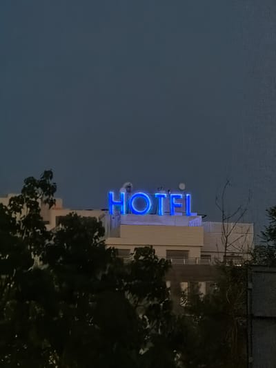 The image captures a dimly lit outdoor scene featuring a hotel sign illuminated with bright blue neon lights, spelling out "HOTEL". The sign is mounted on top of a building, which appears to be a multi-story structure with a light-colored facade and several windows visible. In the foreground and along the sides of the image, the dark silhouettes of trees and branches obscure parts of the building and the sky. The sky above is a deep, muted blue, suggesting it is either dusk or early morning. The overall ambiance is subdued due to the low light conditions.