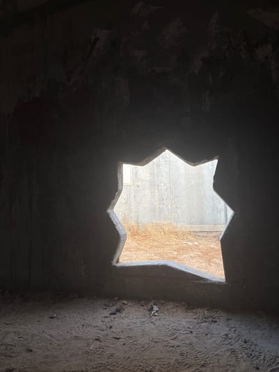 The image depicts the interior of a rough, unfinished concrete structure. A large, jagged opening in the wall offers a view of the exterior. Through this opening, a concrete wall is visible, with dry, brown vegetation in front of it. The floor of the interior is covered in dirt and sand, showing numerous footprints. The lighting is dim inside the structure, with the exterior being brighter. There are no people or animals visible in the image. The scene suggests an abandoned or under-construction site. The mention of Eshkol Regional Council, Israel, implies the location of this structure.