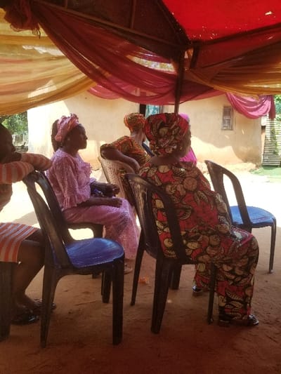 The scene depicts an outdoor gathering in Ochadam, Nigeria, under natural daylight. A temporary canopy, formed by a frame draped with red and yellow translucent fabrics, provides overhead cover. Five individuals are visible, predominantly women, seated on dark-colored plastic chairs on reddish-brown earth. On the far left, a person wearing a white and orange striped garment is partially visible. Next, a woman in a light pink traditional Nigerian garment and matching headwrap faces left. Behind her, a woman in an orange and brown patterned traditional garment and coordinating headwrap faces right. In the immediate foreground, a woman wearing a vibrant red, yellow, and black patterned traditional Nigerian garment and matching headwrap is seated with her back partially to the camera, facing towards the right. Her feet, clad in open-toed sandals, are visible. To the right, another person in pink attire is partially visible. In the background, a light-colored building with a small, dark window opening is present, with vegetation to its left.