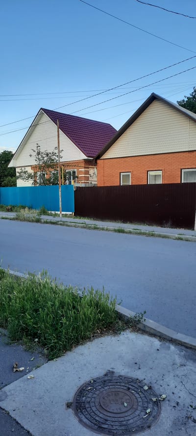 The image depicts a residential street scene in the city of Shakhty, Russia. Two houses with pitched roofs are visible, one with a dark red roof and white siding, and the other with a light beige roof and orange brick facade. Both houses are partially obscured by fences; a bright blue metal fence runs in front of the first house, and a dark brown metal fence separates the second house from the street. The foreground shows a paved road and a sidewalk with green grass and weeds growing along the edge. A circular metal manhole cover is visible on the sidewalk. The sky is clear and blue, suggesting daytime. No people or vehicles are present in the frame.
