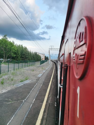 This is a photograph taken from the perspective of being on a moving train at a station. The foreground is dominated by the side of a bright red train car, with a large circular logo and the number "1" visible. The train appears to be departing or arriving at a station, as indicated by the platform and track to the left.

The setting is an outdoor train station with a paved platform and a gravel area beyond a green metal fence. Trees line the edge of the station, suggesting a rural or suburban location. In the distance, a building with a light-colored facade and windows can be seen. Overhead, power lines stretch across the sky, which is partially cloudy with patches of blue.

The time of day appears to be late afternoon or early evening, given the lighting and the quality of the sky. The weather seems to be fair, with a mix of sunlight and clouds. There is no visible activity of people.

The visible text on the train includes Russian lettering within the circular logo, which appears to be "РЖД" (RZD), the abbreviation for Russian Railways. The number "1" is also clearly visible on the train car. The overall impression is one of travel and the infrastructure of a railway system in Russia.