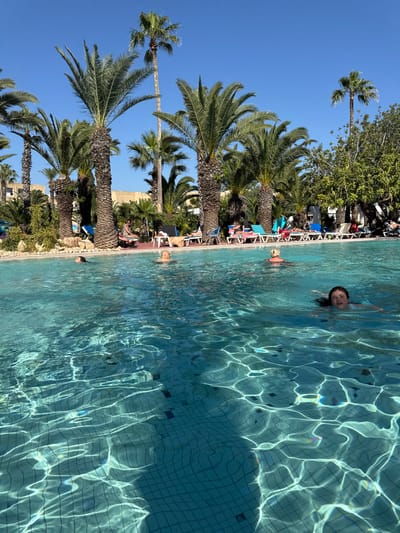 The image shows a swimming pool in Mrezga, Tunisia, surrounded by palm trees and sunbathers. A girl is swimming in the pool, looking directly at the camera. The pool water is light blue, with reflections of sunlight. Lounge chairs and people are visible along the pool's edge. The sky is a clear blue.