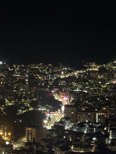 The image is a nighttime, high-angle, wide shot of a densely populated urban area with numerous buildings illuminated by interior lights and streetlights. The cityscape appears to be situated on a hillside, with structures built at varying elevations. The overall scene is dark, with the exception of the artificial lights. In the foreground, a few buildings are clearly visible, including one prominent multi-story structure with illuminated windows. Further into the scene, the lights create a textured mosaic across the landscape, highlighting the density of the urban development. A road with visible lights, presumably from vehicles, snakes through the middle ground of the image. The background fades into darkness, with only distant, faint lights suggesting the continuation of the city or a hazy sky. The image does not contain any people or discernible actions. The environment is exclusively characterized by the built urban infrastructure and the nighttime atmosphere. This image depicts a section of the city of Košljun, Montenegro, at night.