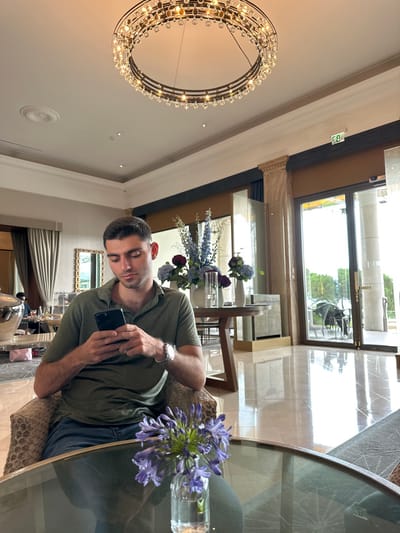 The image depicts the interior of a sophisticated lounge or lobby, specifically identified as the Beau-Rivage Palace Hotel in Lausanne, Switzerland.

In the foreground, a male subject with short, dark hair is seated in a patterned armchair. He is wearing an olive green polo shirt and blue jeans, with a silver-toned watch visible on his left wrist. He is actively engaged with a black smartphone, holding it with both hands and directing his gaze downwards at its screen. In front of him, on a circular glass-top table, a clear glass vase contains a bouquet of purple Agapanthus flowers.

The environment features a highly polished, light-colored marble floor that reflects interior and exterior elements. Overhead, a large, dark-framed circular chandelier adorned with numerous crystal pendants is prominently centered. In the mid-ground, a dark wooden table holds multiple large floral arrangements in vases, displaying a mix of blue and maroon flowers. To the left, a large, reflective silver urn or sculpture is visible, along with another seating area and a mirror, where other individuals are partially obscured. A light pink bag rests on the floor in this area. To the right, large, floor-to-ceiling glass doors or windows provide a view of an outdoor area with trees and outdoor furniture. An illuminated green "EXIT" sign is mounted above these glass doors. The ceiling is white, featuring decorative molding and recessed lighting, indicating an elegant and well-maintained space. The scene is brightly lit, consistent with daytime.