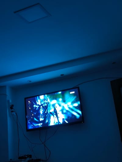 In a dimly lit room with blue ambient lighting, a flat-screen television is mounted on a wall. The TV displays a close-up image of a metallic, skull-like face with glowing blue eyes, possibly from a movie or video game. Various cables are connected to the TV and run down to a shelf below, where electronic devices are placed. A small camera is mounted on the wall near the TV. The room appears to be a residential setting.