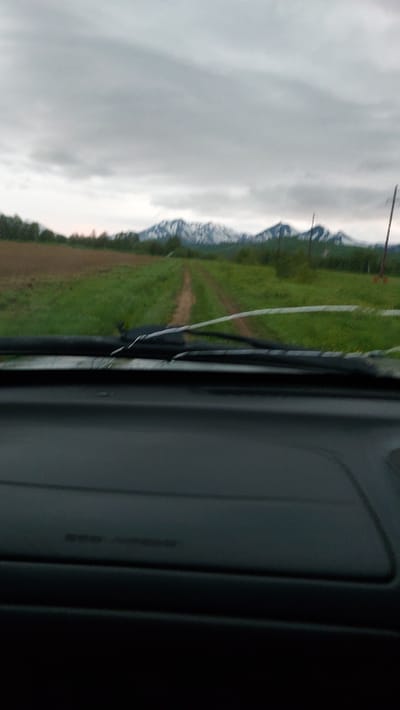 The image depicts a view from inside a vehicle, looking out through the windshield. The dashboard is visible in the foreground. Outside the vehicle, a dirt road extends into a grassy field, leading towards snow-capped mountains in the distance under a cloudy sky. Power lines are also visible on the right side of the frame. The scene appears to be a rural landscape. The location is not specified in the image itself, and therefore cannot be identified as Lesnoi, Russia.