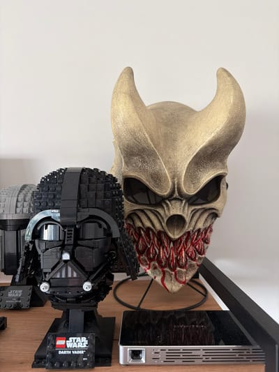 The image shows a collection of items on a wooden shelf against a white background, possibly in an apartment in Малое Седельниково, Russia. In the foreground and slightly to the left is a Lego build of Darth Vader's helmet, displayed on a black stand with a "LEGO STAR WARS DARTH VADER" plaque. To the right of the Darth Vader helmet, and slightly behind it, is a grotesque mask with golden horns, dark eye sockets, and a mouth filled with bloody teeth. The mask is mounted on a black stand. To the left of the Darth Vader helmet, a portion of another Lego build, possibly a helmet, is visible, rendered in gray bricks. In the lower right of the image, a silver electronic device with ventilation slots and a port is partially visible. The lighting suggests an indoor setting. There are no people or animals present in the image.